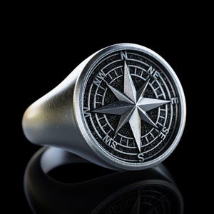 May include: A silver-colored ring with a compass rose design. The ring features a raised compass rose with directional markers and a star in the center. The ring is set against a black background.
