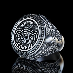 May include: A silver ring with a detailed design featuring a scorpion and the words "Memento Mori" and "Scorpio".
