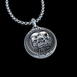 May include: Silver pendant necklace with a circular design featuring two skulls with a single eye and the text "Memento Mori Gemini".