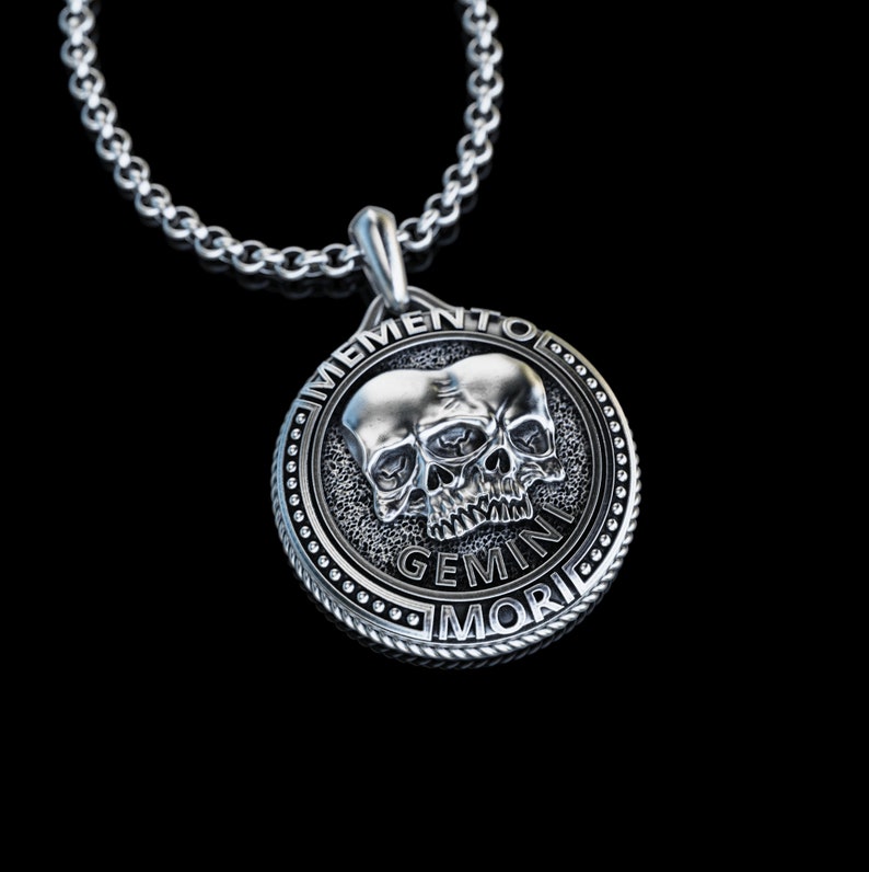 May include: Silver pendant necklace with a circular design featuring two skulls with a single eye and the text "Memento Mori Gemini".