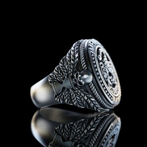 May include: A silver ring with a detailed design featuring a skull and a rope-like pattern. The ring has a raised circular section with a detailed design.