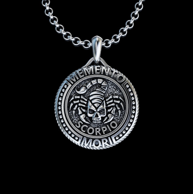 May include: Silver pendant necklace with a round charm featuring a skull with scorpion wings and the text "Memento Mori Scorpio".