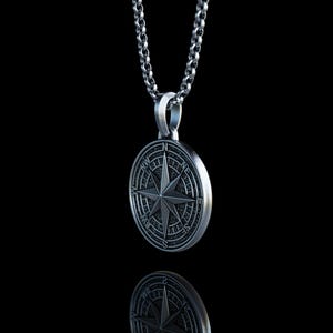 May include: A silver-colored compass pendant with a star design, hanging from a matching chain. The pendant is round with engraved details, reflecting on a black surface. The necklace is a classic accessory.