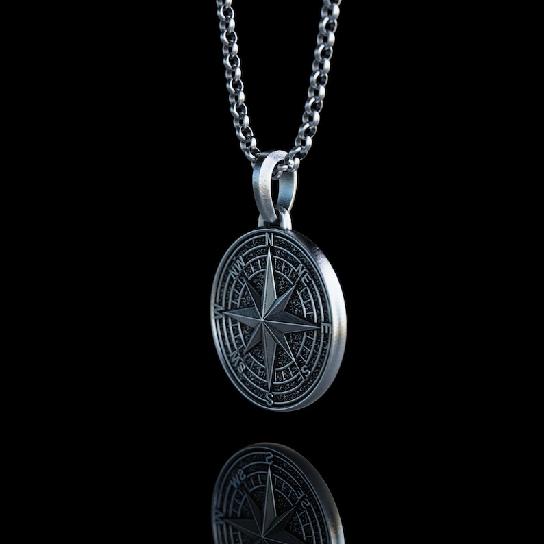 May include: A silver-colored compass pendant with a star design, hanging from a matching chain. The pendant is round with engraved details, reflecting on a black surface. The necklace is a classic accessory.