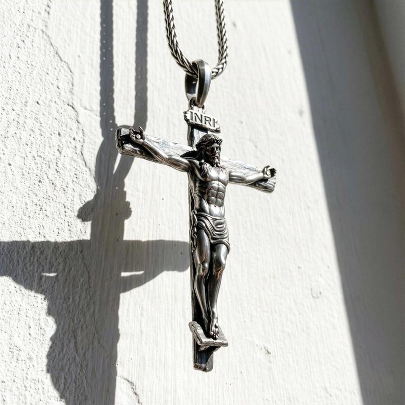 Puede incluir: Un collar con colgante plateado que presenta un crucifijo detallado. El crucifijo representa a Jesucristo en la cruz, con la inscripci&oacute;n "INRI" en la parte superior. El colgante cuelga de una cadena plateada, sobre una pared blanca texturizada.