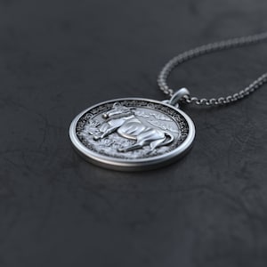 May include: A silver pendant necklace with a round charm featuring a detailed 3D image of an ox or bull. The charm has a textured border and a detailed background.