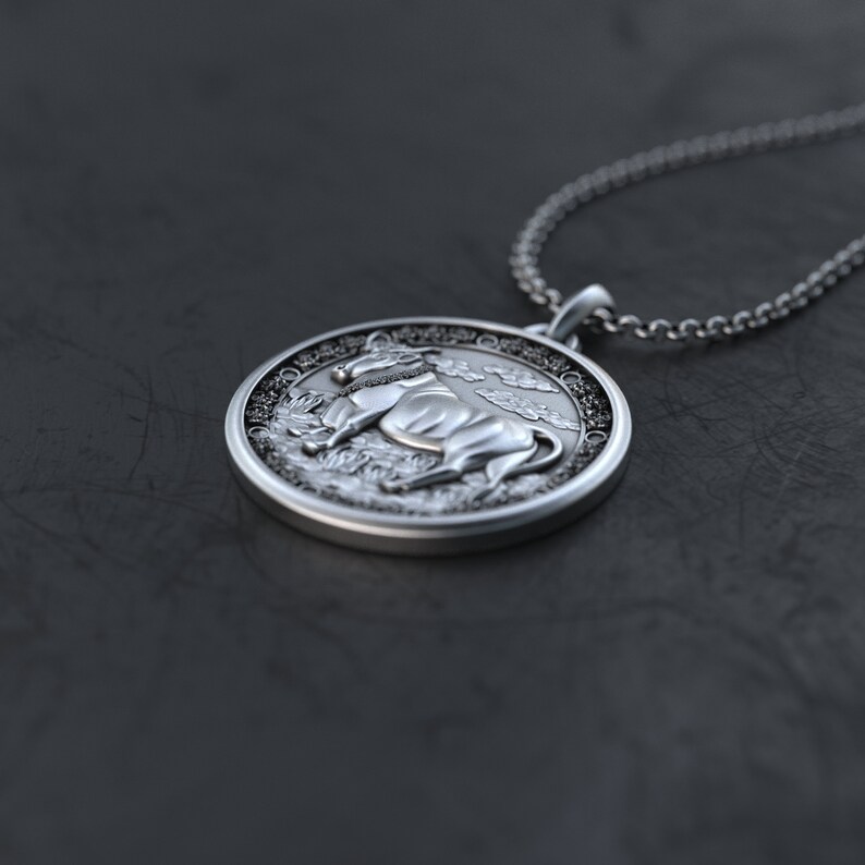 May include: A silver pendant necklace with a round charm featuring a detailed 3D image of an ox or bull. The charm has a textured border and a detailed background.