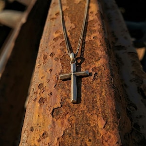 Puede incluir: Un colgante de cruz plateada cuelga de una delicada cadena. La cruz tiene un dise&ntilde;o geom&eacute;trico simple. El collar se muestra sobre una superficie met&aacute;lica marr&oacute;n oxidada y texturizada.