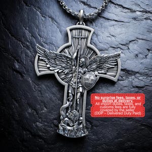 Op de afbeelding: Een zilverkleurige hangerketting met een gedetailleerd kruisontwerp. De hanger toont een engel met vleugels, die een speer en schild vasthoudt. Het schild heeft de tekst "QUIS UT DEUS". De ketting ligt op een donkere, getextureerde ondergrond.