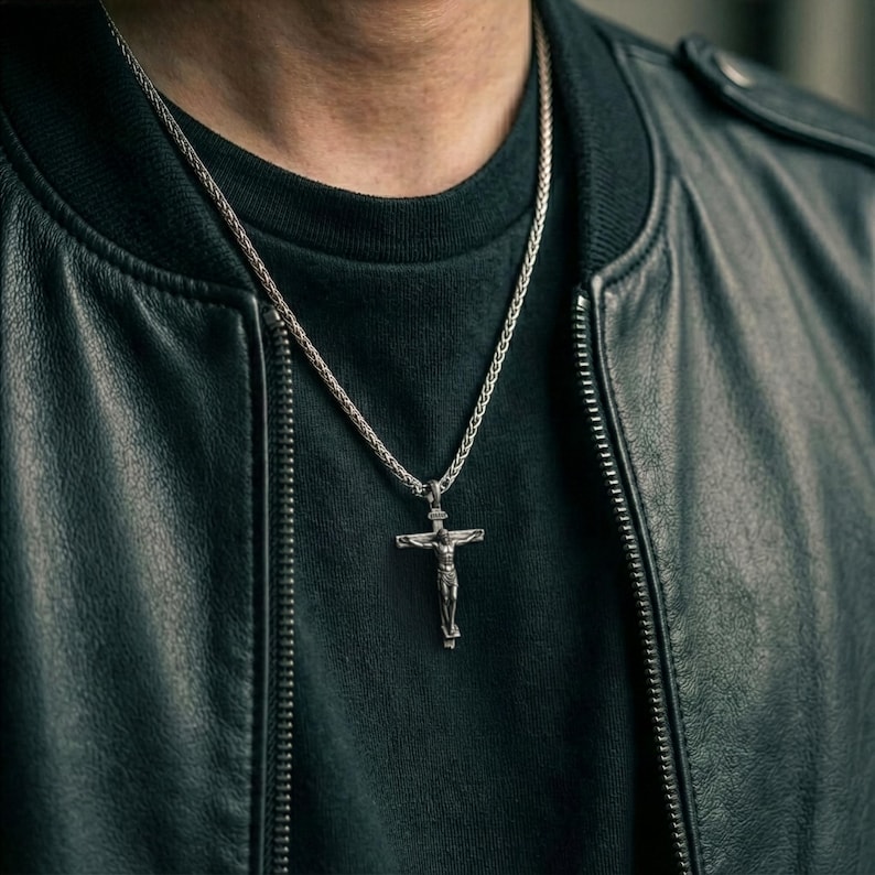 Puede incluir: Collar con colgante de crucifijo plateado. El colgante representa una figura en una cruz. El collar se lleva sobre una camisa negra y una chaqueta de cuero negra.