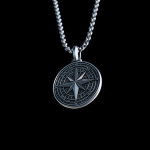 May include: A silver-colored compass pendant with a detailed design, suspended from a matching chain. The pendant features a prominent star in the center, surrounded by directional markings. The background is a solid black.