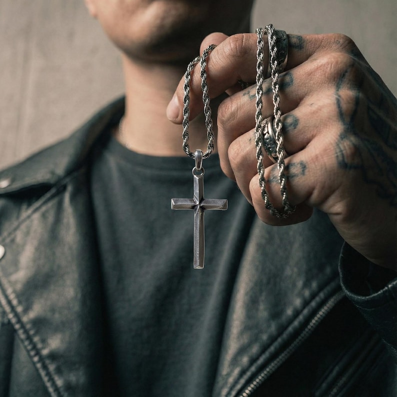 Puede incluir: Collar con colgante de cruz plateada y cadena de cuerda. La cruz tiene un dise&ntilde;o simple y minimalista. El collar se sostiene sobre un fondo oscuro, mostrando los detalles de la joya.