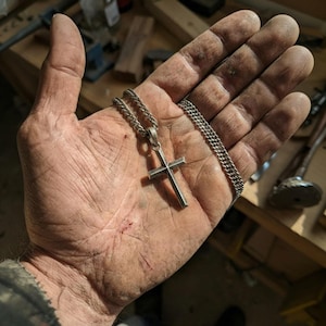 Puede incluir: Un collar con colgante de cruz plateada y cadena de eslabones. La cruz tiene un dise&ntilde;o simple y minimalista. El collar se muestra en una mano con suciedad y desgaste visibles, lo que sugiere un taller o una obra.
