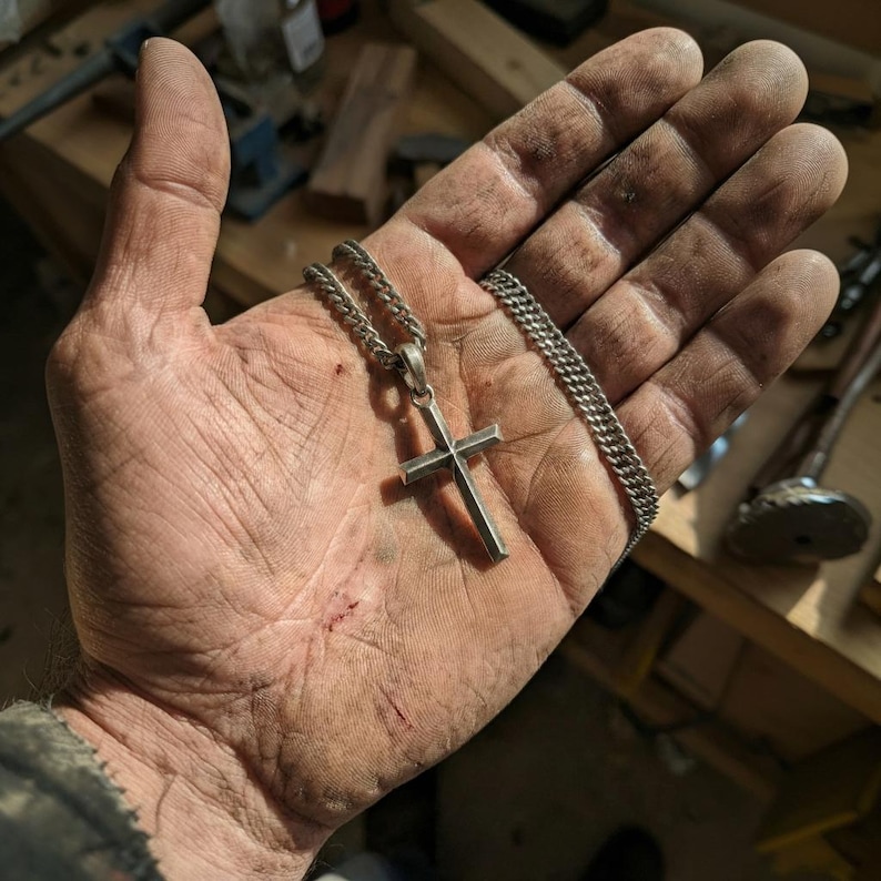 Puede incluir: Un collar con colgante de cruz plateada y cadena de eslabones. La cruz tiene un dise&ntilde;o simple y minimalista. El collar se muestra en una mano con suciedad y desgaste visibles, lo que sugiere un taller o una obra.