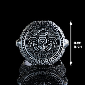 May include: A silver ring with a black scorpion and skull design. The ring is engraved with the words "Memento Mori" and "Scorpio". The ring is 0.85 inches in diameter.