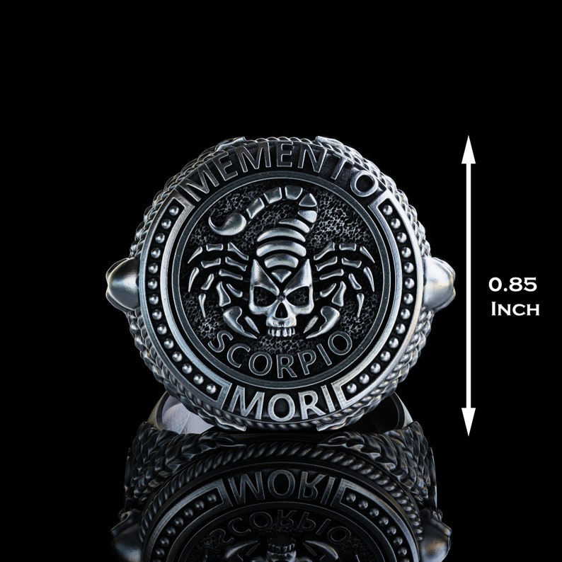 May include: A silver ring with a black scorpion and skull design. The ring is engraved with the words "Memento Mori" and "Scorpio". The ring is 0.85 inches in diameter.