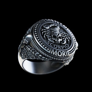 May include: A silver ring with a detailed design featuring a scorpion with a skull and the words "Memento Mori" and "Scorpio".