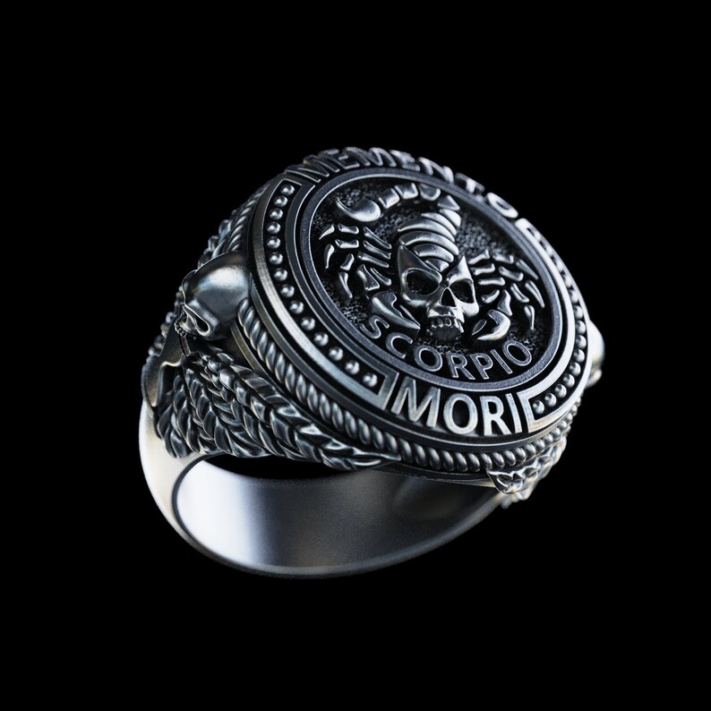 May include: A silver ring with a detailed design featuring a scorpion with a skull and the words "Memento Mori" and "Scorpio".