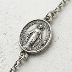 Puede incluir: Primer plano de un collar plateado con un colgante ovalado. El colgante tiene una imagen grabada de una figura religiosa y está inscrito con texto y el año 1830. La cadena está hecha de pequeños eslabones interconectados.