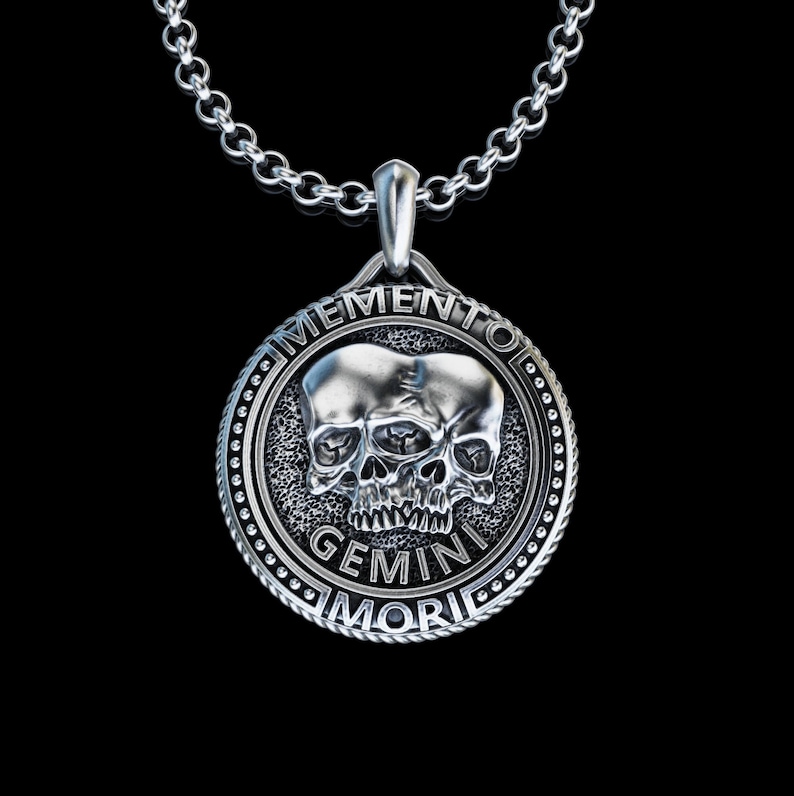 May include: Silver pendant necklace with a circular design featuring two conjoined skulls and the text "Memento Mori Gemini".