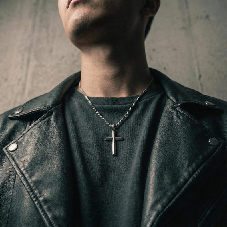 Puede incluir: Collar con colgante de cruz plateada. La cruz cuelga de una cadena fina. El collar se lleva sobre una camiseta gris oscuro y una chaqueta de cuero negra.