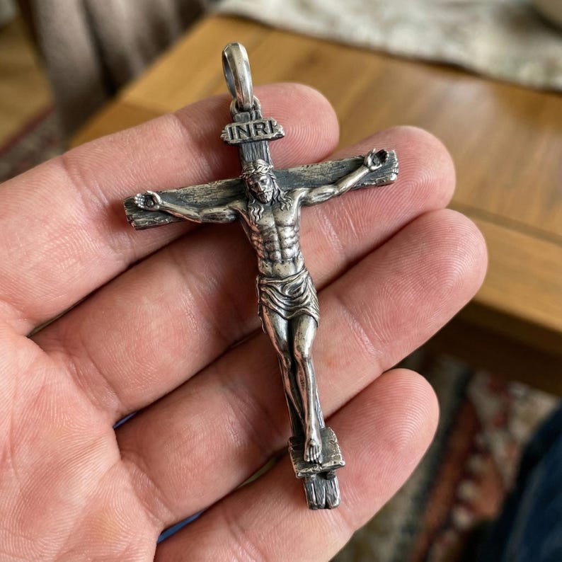 Puede incluir: Un colgante de crucifijo plateado que representa a Jesucristo en la cruz, con la inscripci&oacute;n "INRI" en la parte superior. El colgante se sostiene en una mano, destacando los detalles del s&iacute;mbolo religioso. El colgante es de plata.
