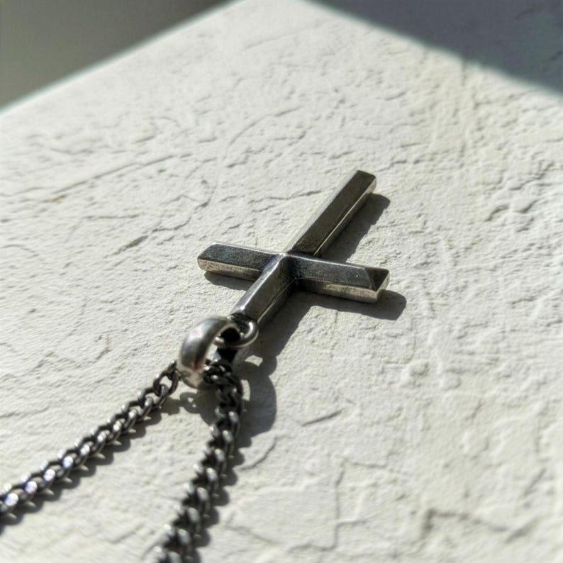 Puede incluir: Un colgante de cruz plateada con una cadena a juego. La cruz tiene un dise&ntilde;o simple y minimalista con l&iacute;neas limpias. La cadena es de estilo curb, y el colgante est&aacute; unido con un peque&ntilde;o lazo. El fondo es una superficie texturizada de color blanquecino.