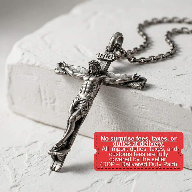 May include: A silver-toned crucifix pendant with a detailed depiction of Jesus on the cross. The pendant hangs from a chain. The word "INRI" is above the cross. The image is set against a textured white background.