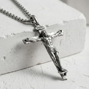 Puede incluir: Un collar con colgante plateado que presenta un crucifijo detallado. El crucifijo representa a Jesucristo en la cruz, con las letras "INRI" sobre su cabeza. El collar tiene una cadena con un dise&ntilde;o de eslabones texturizados.