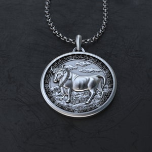 May include: Silver pendant necklace featuring a detailed bull design with a textured background. The bull is depicted standing on a field of grass with clouds in the background.