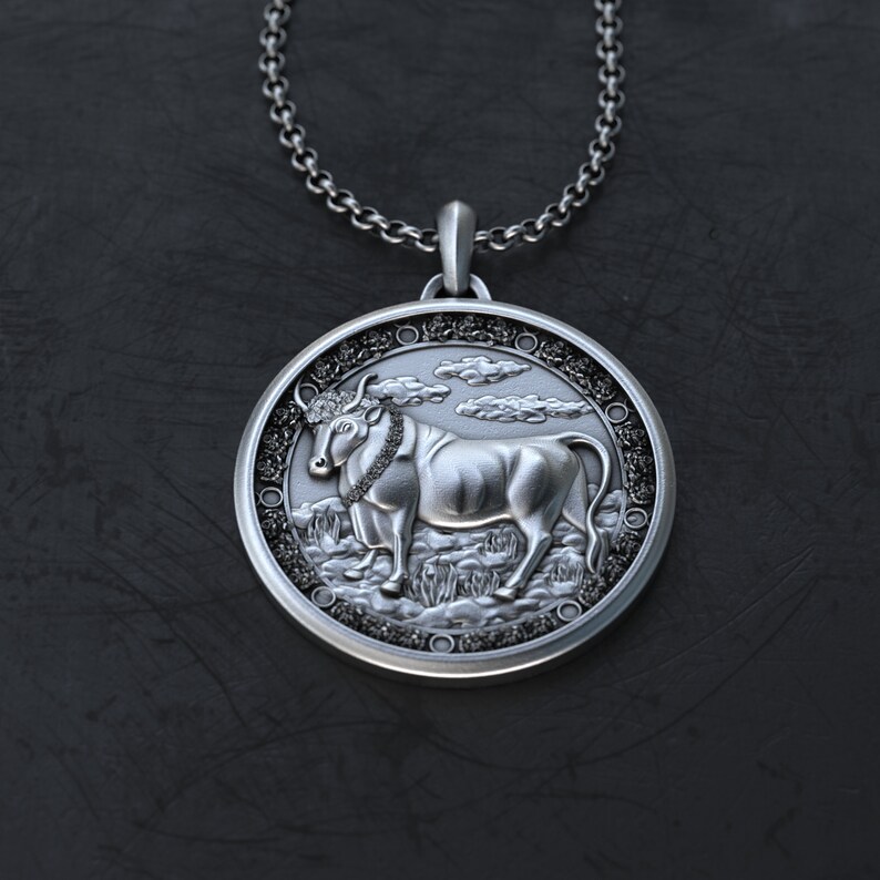 May include: Silver pendant necklace featuring a detailed bull design with a textured background. The bull is depicted standing on a field of grass with clouds in the background.