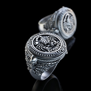 May include: A silver ring with a detailed design featuring a scorpion and skull, surrounded by the words "Memento Mori" and "Scorpio".