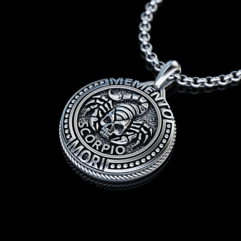 May include: Silver pendant necklace with a round, black, engraved design featuring a scorpion and skull with the words "Memento Mori" and "Scorpio" around the edge.