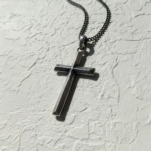 Puede incluir: Collar con colgante de cruz plateada. La cruz tiene un dise&ntilde;o geom&eacute;trico simple con una superficie ligeramente texturizada. La cadena es de estilo oscuro y eslabonado. El collar se muestra sobre un fondo texturizado blanquecino.
