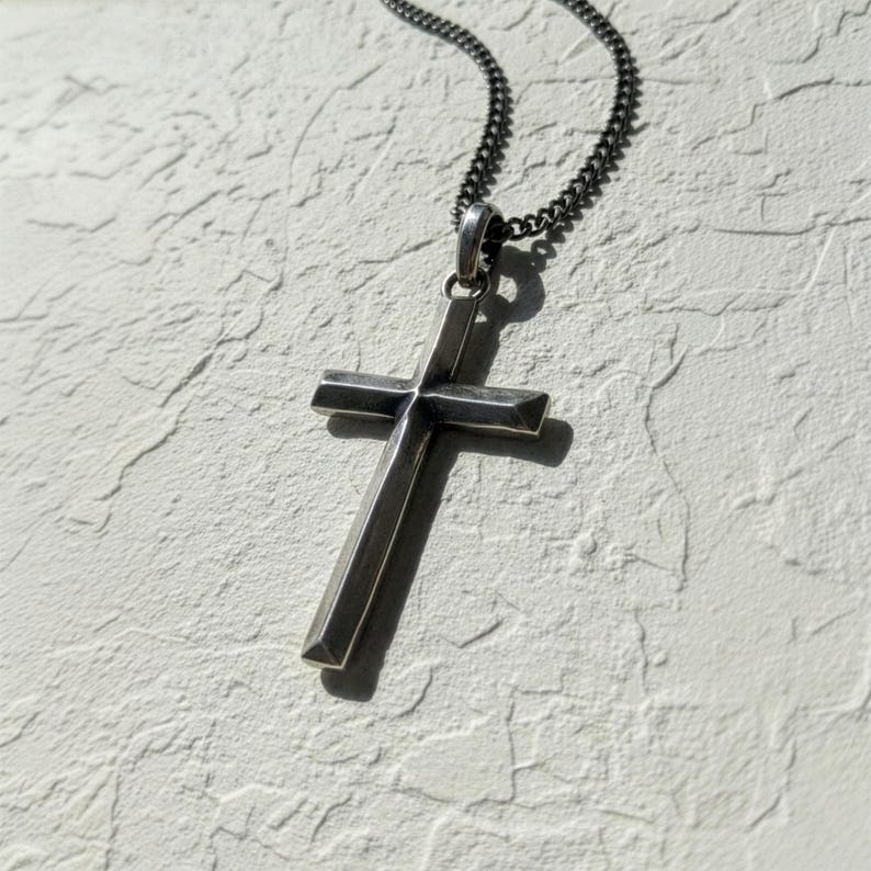 Puede incluir: Collar con colgante de cruz plateada. La cruz tiene un dise&ntilde;o geom&eacute;trico simple con una superficie ligeramente texturizada. La cadena es de estilo oscuro y eslabonado. El collar se muestra sobre un fondo texturizado blanquecino.