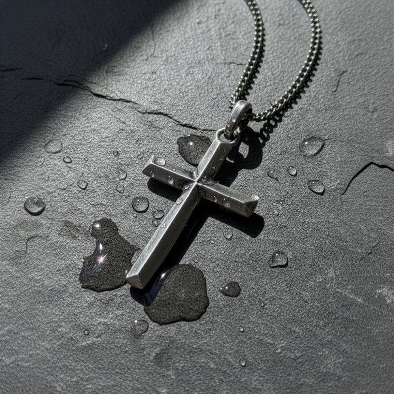 Puede incluir: Un collar con colgante de cruz plateada y cadena oscura. La cruz tiene una superficie texturizada y se muestra sobre una superficie oscura y texturizada con gotas de agua.