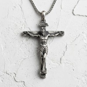Puede incluir: Un colgante de crucifijo plateado con una detallada representaci&oacute;n de Jesucristo en la cruz. El colgante cuelga de una cadena plateada. La cruz tiene las letras INRI en la parte superior.