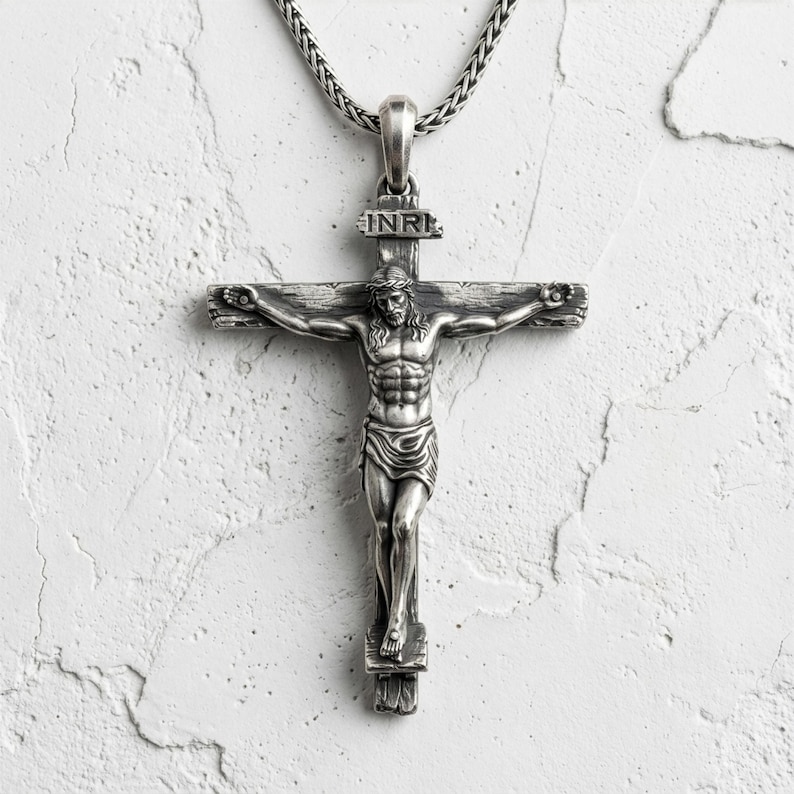 Puede incluir: Un colgante de crucifijo plateado con una detallada representaci&oacute;n de Jesucristo en la cruz. El colgante cuelga de una cadena plateada. La cruz tiene las letras INRI en la parte superior.