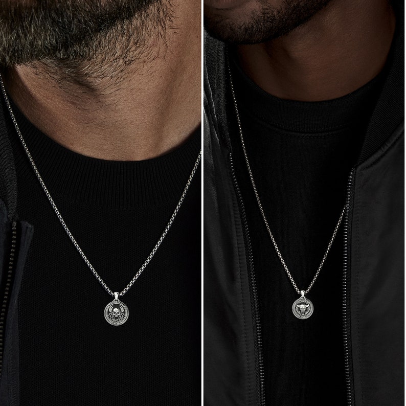 May include: A silver chain necklace with a round pendant featuring a skull design. The pendant is worn by a person wearing a black shirt.