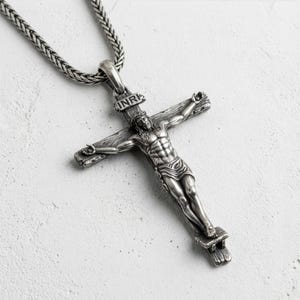 Puede incluir: Un colgante de crucifijo plateado con una detallada representaci&oacute;n de Jesucristo en la cruz. El colgante cuelga de una cadena texturizada. La cruz presenta la inscripci&oacute;n "INRI" en la parte superior.