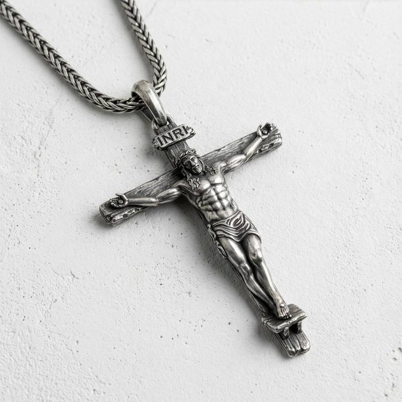 Puede incluir: Un colgante de crucifijo plateado con una detallada representaci&oacute;n de Jesucristo en la cruz. El colgante cuelga de una cadena texturizada. La cruz presenta la inscripci&oacute;n "INRI" en la parte superior.