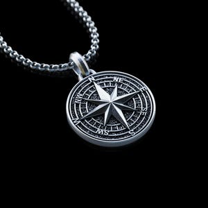 May include: A silver compass pendant necklace. The pendant features a detailed compass rose design with directional markers. The necklace has a chain with a textured, linked design. The background is black.