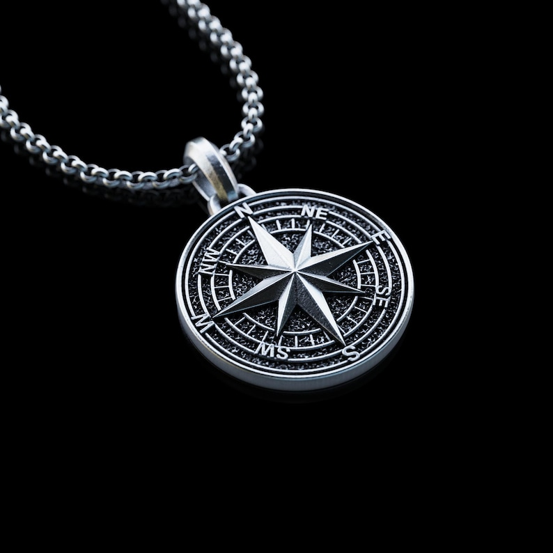 May include: A silver compass pendant necklace. The pendant features a detailed compass rose design with directional markers. The necklace has a chain with a textured, linked design. The background is black.