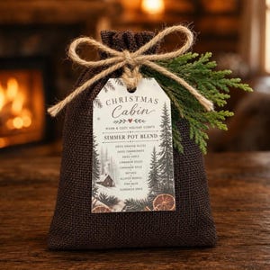 May include: A dark brown burlap drawstring pouch tied with a jute twine bow. A white rectangular tag is attached, featuring the text "CHRISTMAS CABIN WARM & COZY HOLIDAY SCENTS SIMMER POT BLEND" and a list of ingredients, accented by a green evergreen sprig.