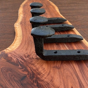 May include: A wall-mounted rack featuring four black metal railroad spikes attached to a piece of natural live-edge wood with a prominent reddish-brown grain pattern. The spikes are arranged in a vertical row, each with two visible screw holes.