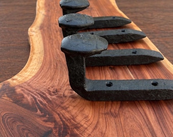 May include: A wall-mounted rack featuring four black metal railroad spikes attached to a piece of natural live-edge wood with a prominent reddish-brown grain pattern. The spikes are arranged in a vertical row, each with two visible screw holes.