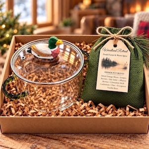 May include: A clear glass pot with green handles and a lid topped with a carved wooden mallard duck figure. Beside it sits a green burlap bag with a tag labeled Woodland Retreat Fresh Forest & Warm Spice Simmer Pot Blend with ingredients.