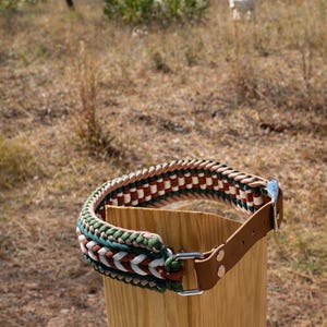 May include: A handcrafted dog collar with a braided design in green, white, brown, and red. The collar features a brown leather strap and a silver-colored buckle. The collar is displayed on a wooden post outdoors.