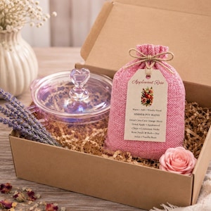 May include: A gift box contains a clear glass pot with a heart-shaped handle, a pink sachet labeled "Applewood Rose Simmer Pot Blend", a bundle of lavender, and a pink rose. The box is filled with shredded paper.