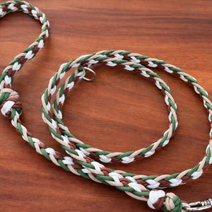 May include: Braided dog leash in shades of green, brown, white, and tan. The leash features a loop handle and a metal clasp. The leash is lying on a wooden surface.